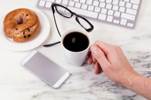 Overhead view marble counter top with male hand holding cup of coffee with computer keyboard, cell phone, reading glasses, and bagel on plate. Work at home concept.